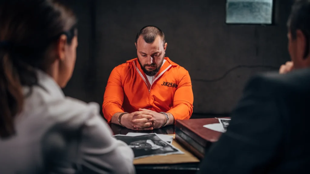 Man in an orange prison uniform sitting at a table during an interrogation.