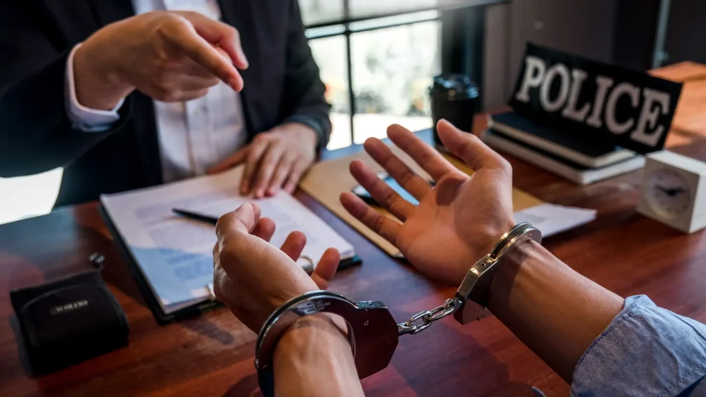 Person in handcuffs sitting at a desk while talking to someone pointing at documents.